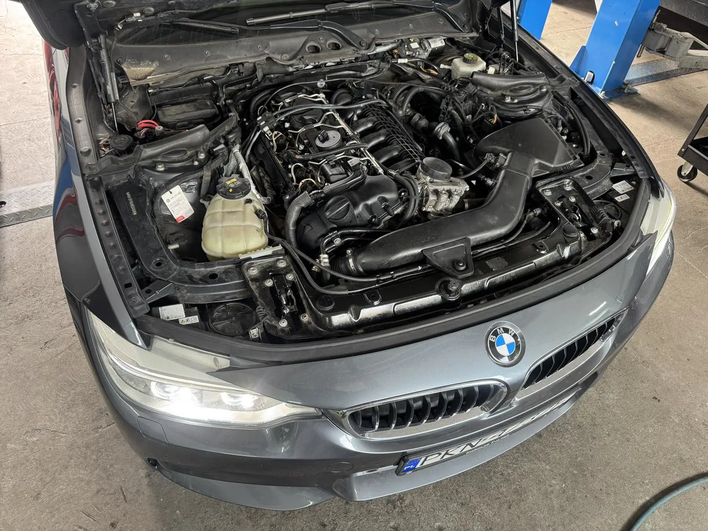 Szary BMW w warsztacie, otwarta maska i widoczny silnik podczas przeglądu.
